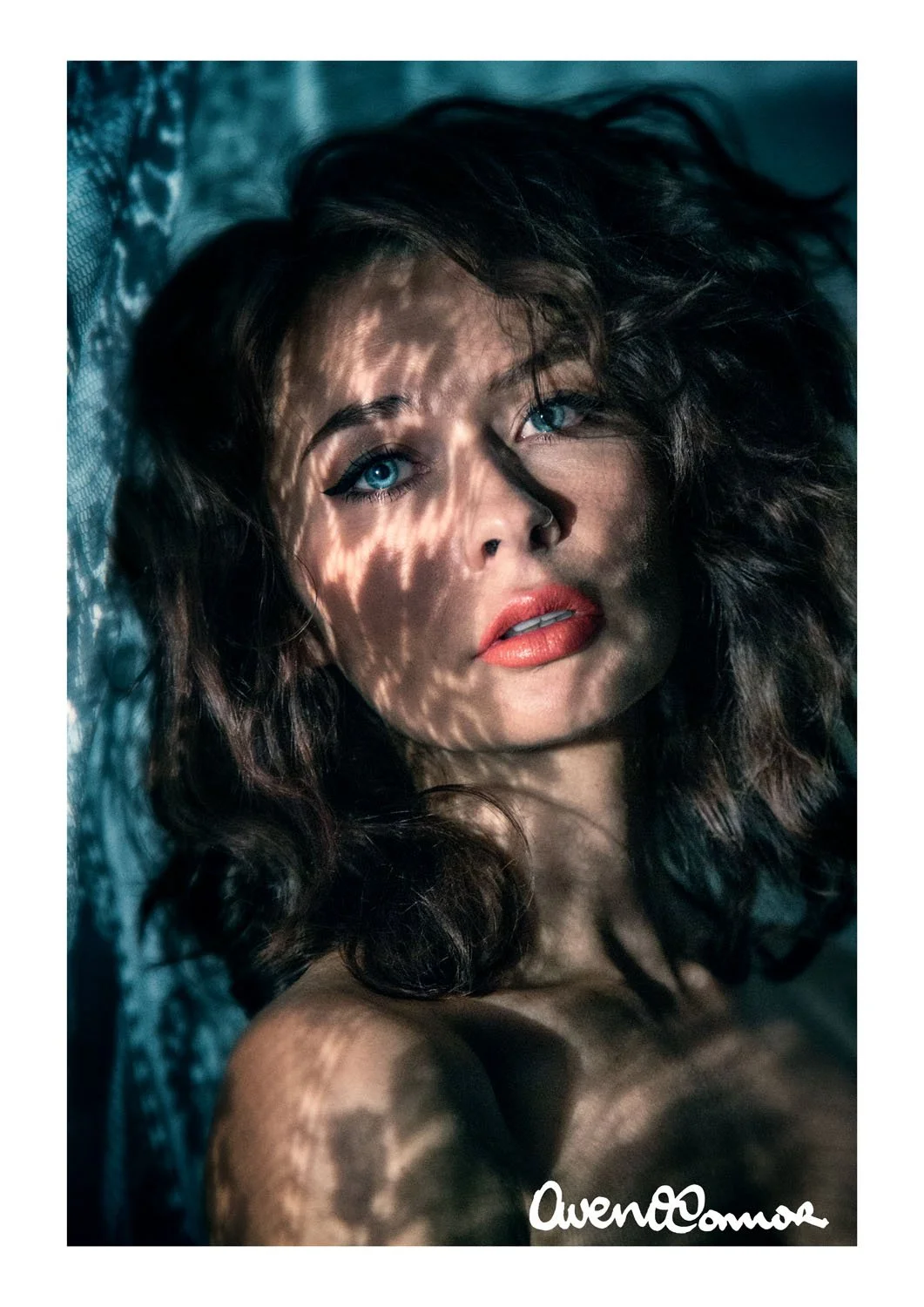Close-up of a woman with blue eyes, dark wavy hair, lips slightly parted, lying on a textured surface with dappled sunlight casting shadows on her face.