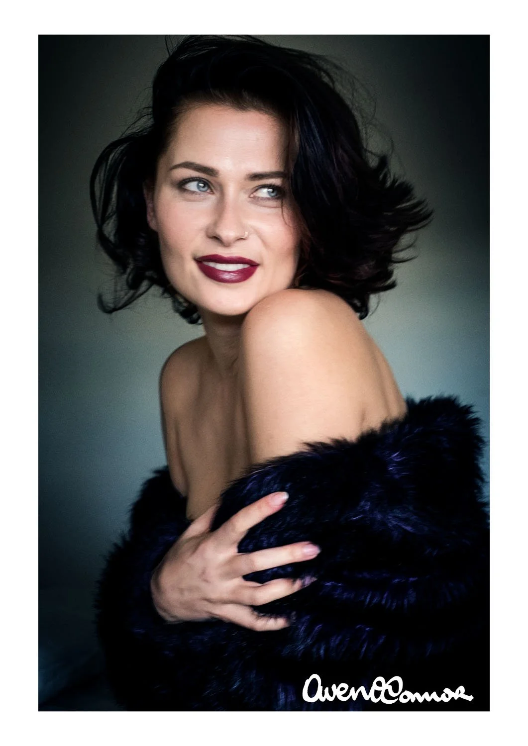 Portrait of a woman with short dark hair, wearing dark lipstick, and a black faux fur stole, looking to the side with a slight smile, against a plain background.