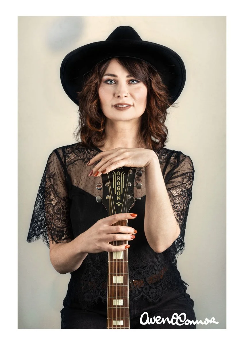 A woman with wavy brown hair wearing a black hat and lace black top, holding a guitar vertically in front of her, with her left hand resting on top of the guitar neck. She has makeup with brown lipstick and is looking at the camera. The background is