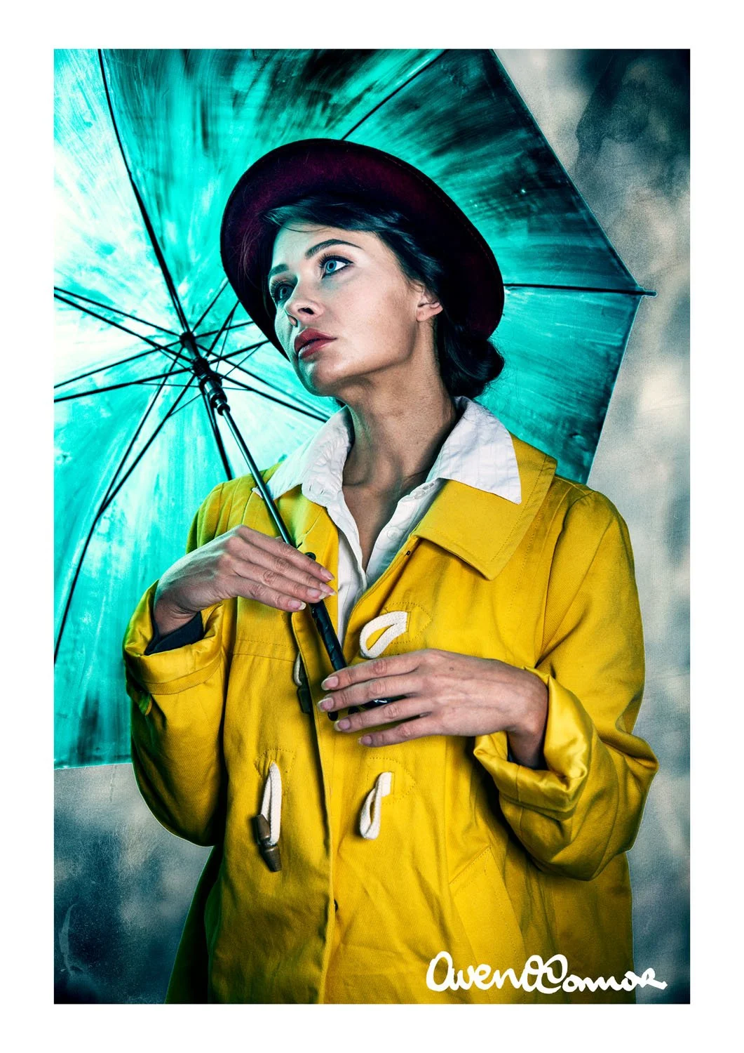A woman with blue eyes and dark hair wearing a yellow raincoat, white shirt, and a dark hat, holding a blue umbrella with rain in the background.