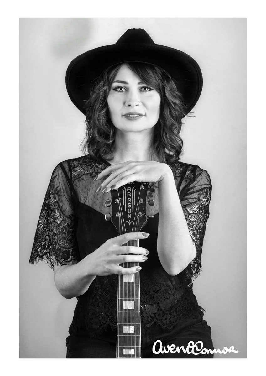 A woman with wavy hair wearing a wide-brimmed hat and lace top, holding an electric guitar, in a black and white portrait.