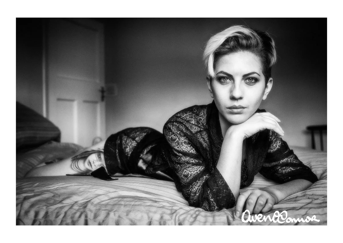 Black and white photo of a woman with short blonde hair lying on a bed, resting her chin on her hand, with a serious expression, wearing a lace top. There is a tattoo of a woman's face on her arm. The room has a closed white door in the background.