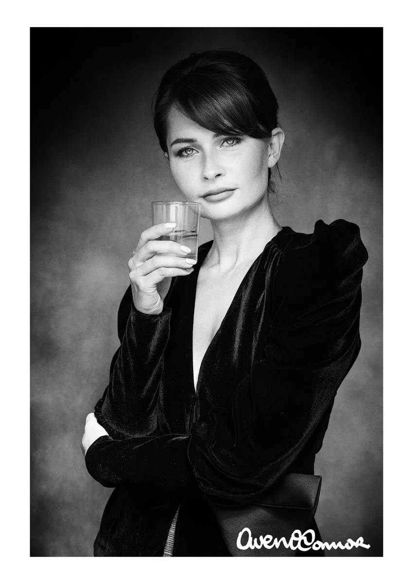 Black and white portrait of a woman with short dark hair, holding a glass of water, wearing a velvet long-sleeve top, with a confident expression and signature at the bottom right.