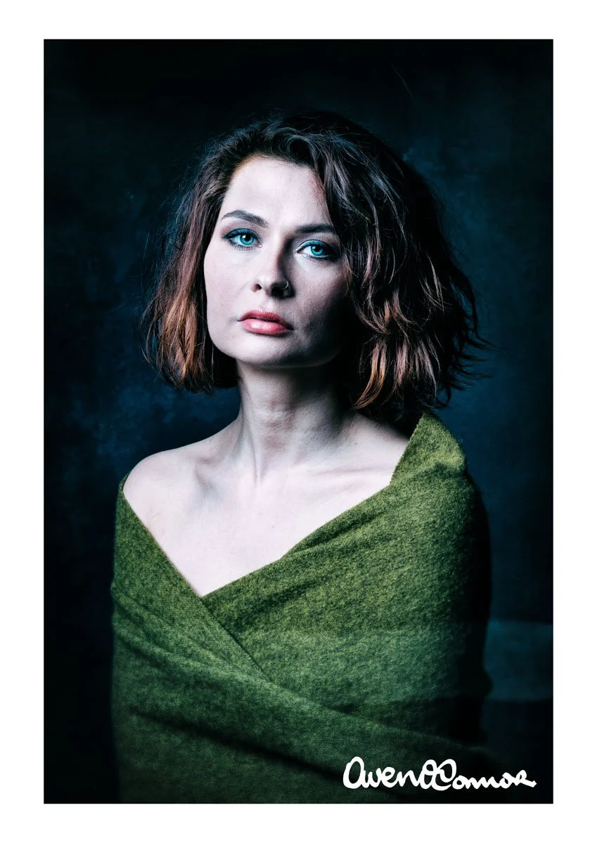 Portrait of a woman with shoulder-length wavy brown hair, blue eyes, and light makeup, wrapped in a green shawl against a dark background.