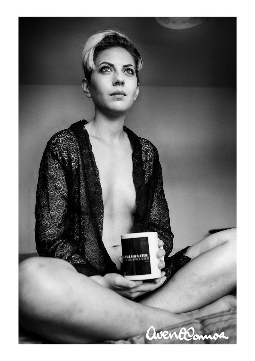 Black and white photo of a woman with short hair, sitting cross-legged, holding a mug, wearing a lace robe, looking upward with a thoughtful expression.