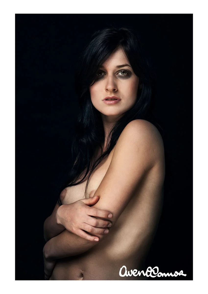 A woman with long black hair and fair skin poses topless against a black background, crossing her arms over her chest, with an intense gaze at the camera.