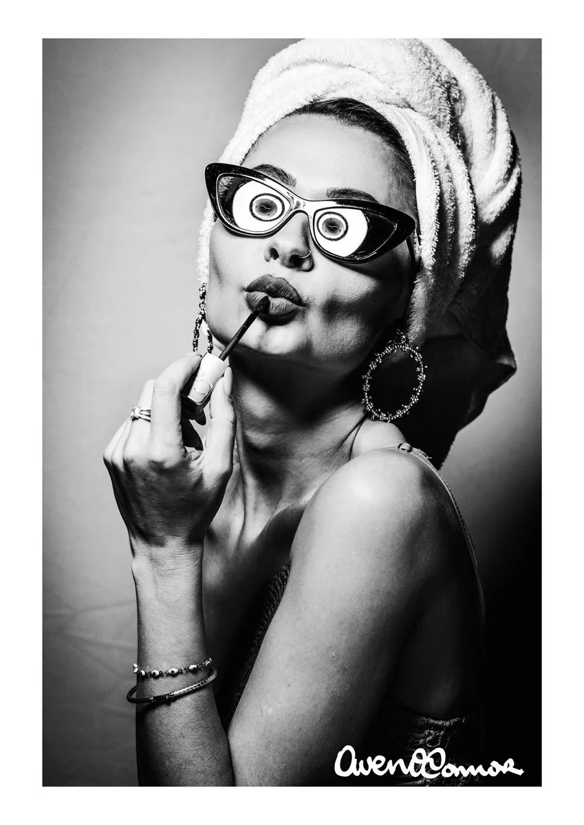 Black and white portrait of a woman with a towel wrapped around her head, wearing large unique sunglasses, hoop earrings, and bracelets, holding a lipstick applicator near her lips, signed by Avenue Omore.