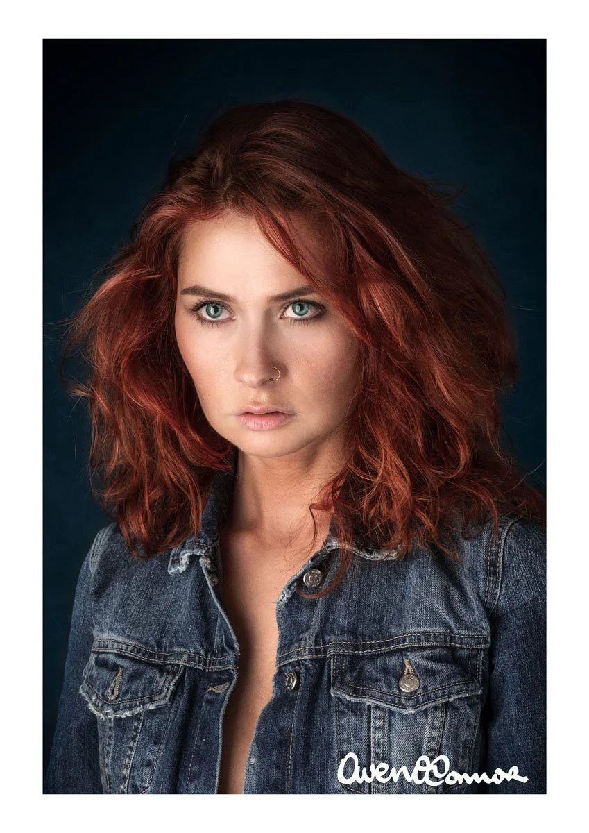 A young woman with red, curly hair and blue eyes wearing a denim jacket against a dark blue background.