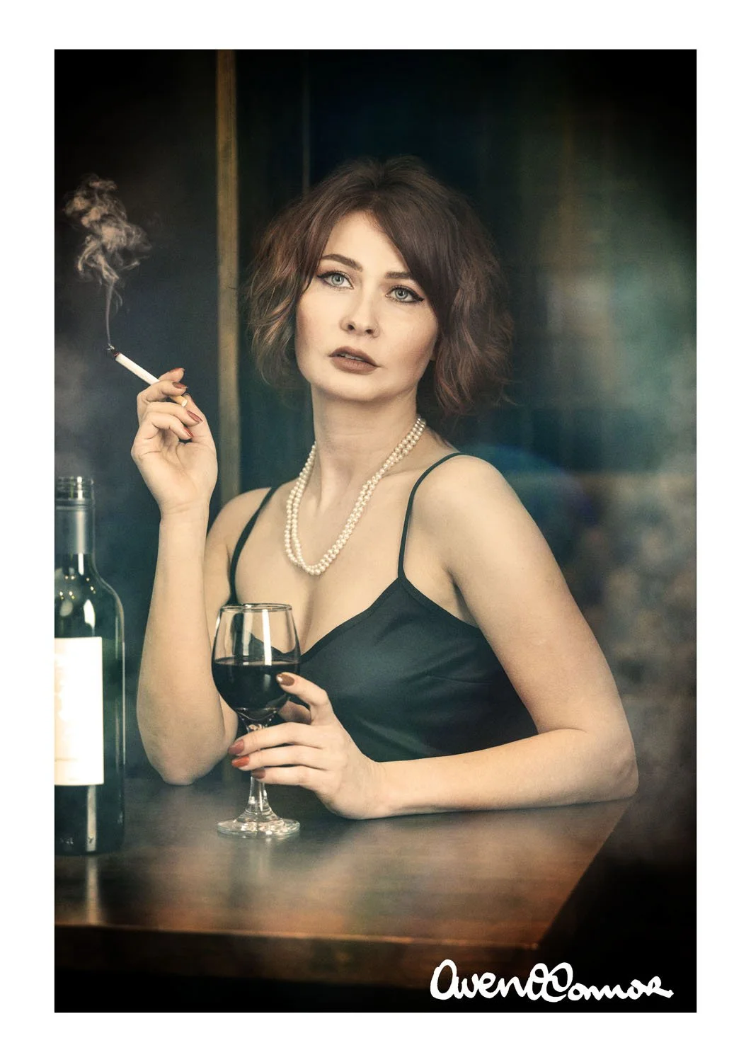 A woman with short, wavy brown hair, wearing a black spaghetti strap dress and pearl necklace, is holding a glass of red wine in one hand and a lit cigarette in the other, standing beside a bottle of wine, with a dark background.