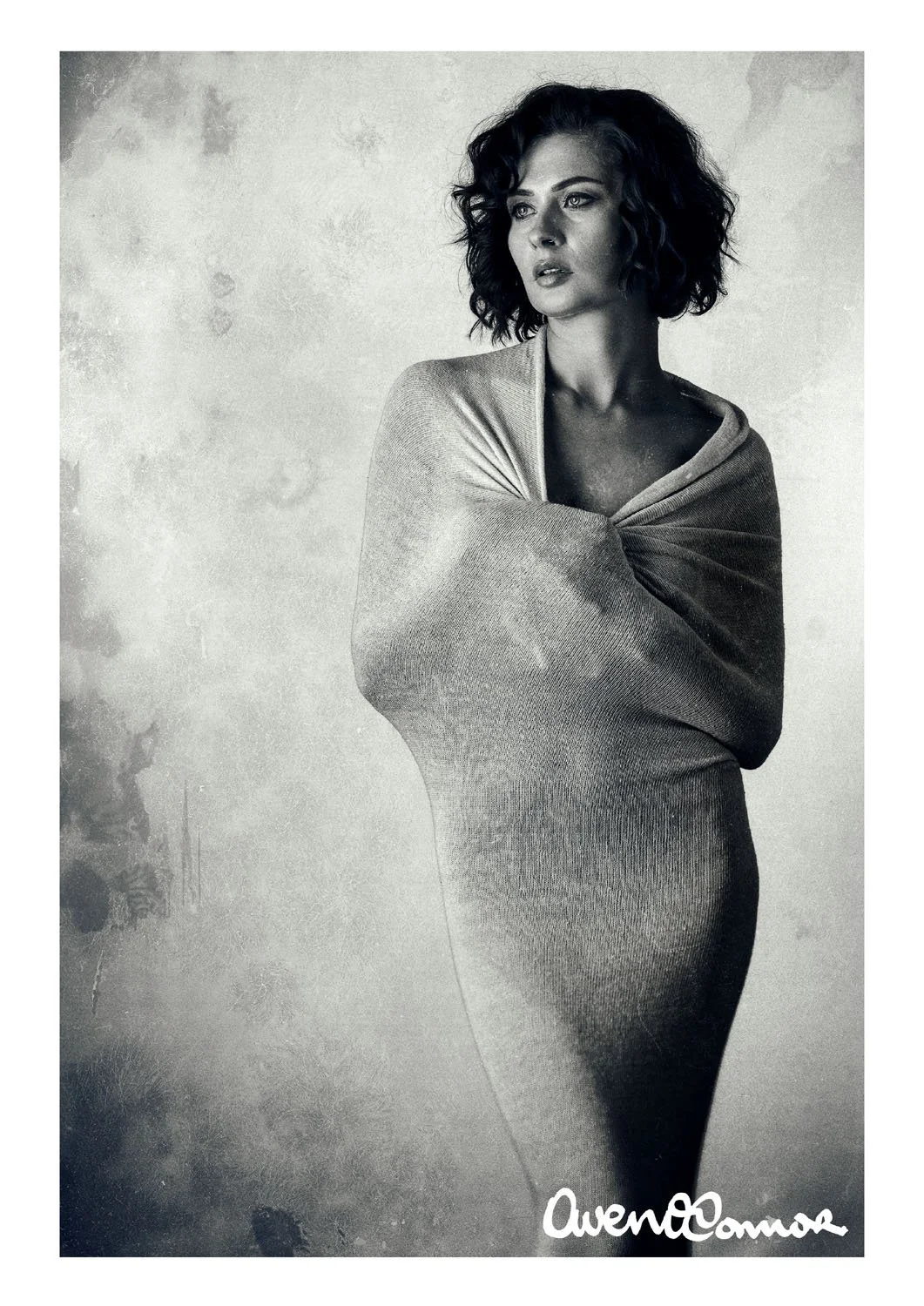 Black and white portrait of a woman with curly hair, wearing a long dress and a wrap or shawl, standing with her arms crossed against a textured background