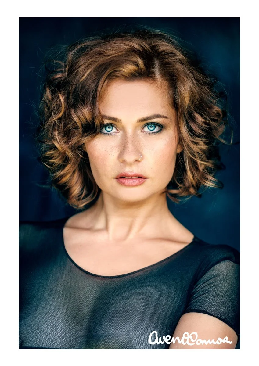 Close-up portrait of a woman with bright blue eyes, short curly brown hair, wearing a black sheer top, with a deep blue background.