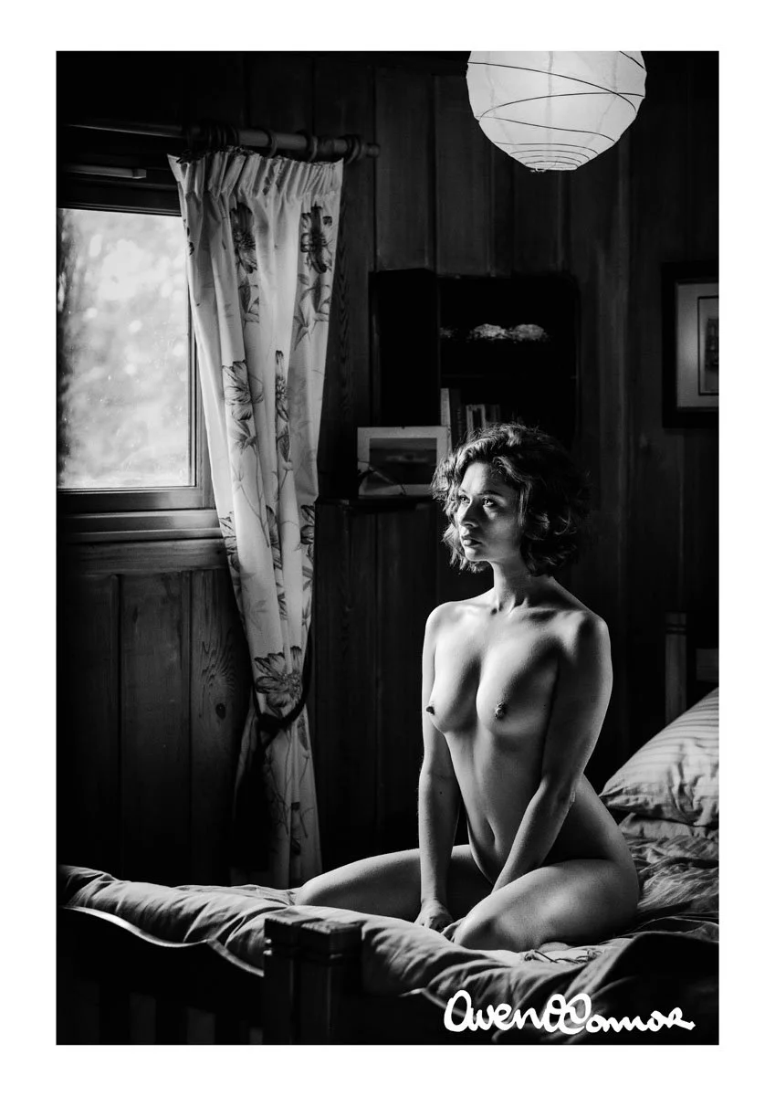 A black and white photo of a nude woman with curly hair, kneeling on a bed in a wooden-paneled room, looking out the window to the left. The room has a curtain with a floral pattern, a bookshelf with books, and a hanging paper lantern. The signature