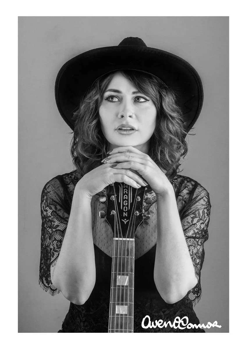 Black and white portrait of a woman wearing a large hat, lace top, and holding a guitar with the name 'Dragon' on it, with her hands clasped under her chin.