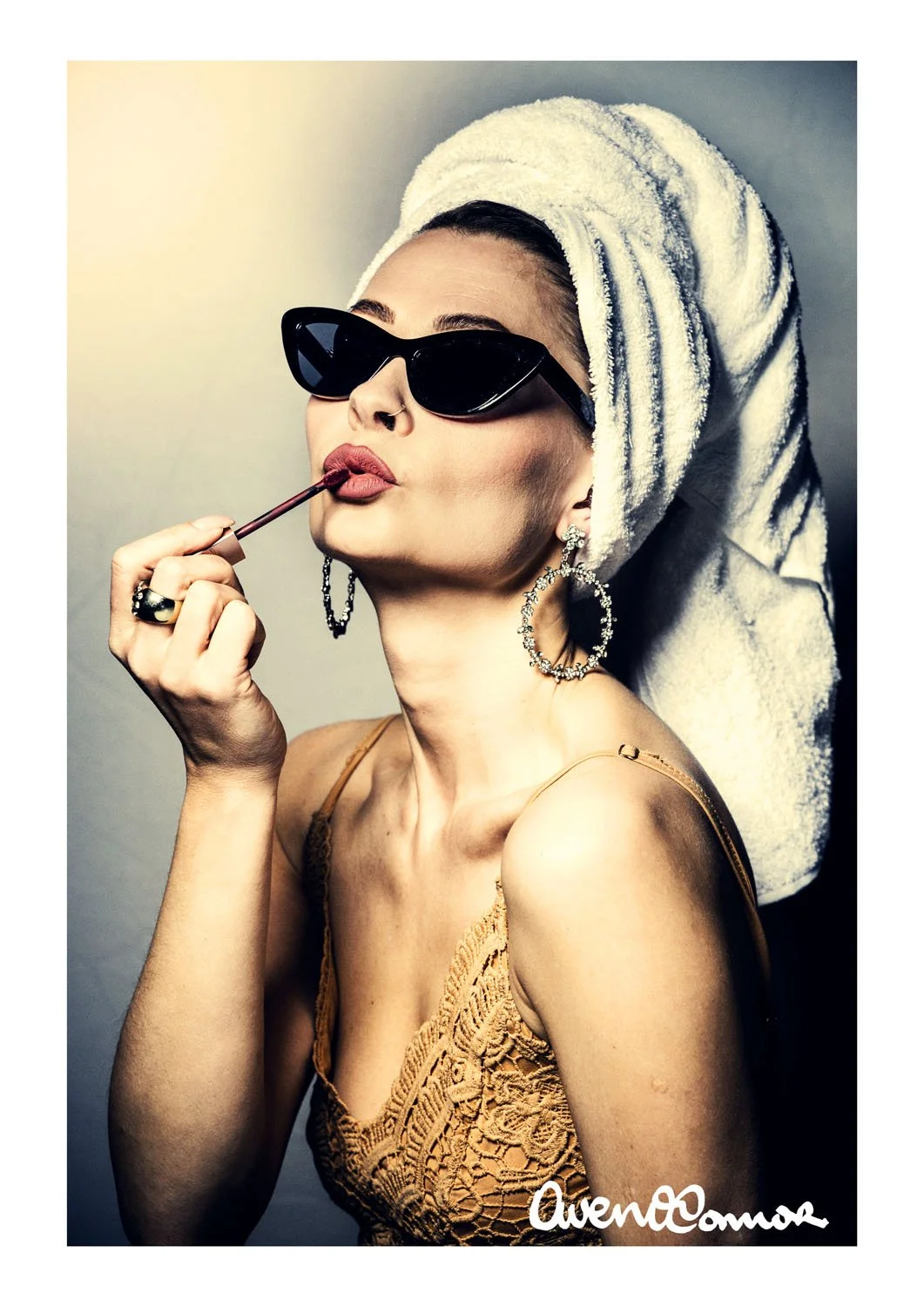 A woman with a towel wrapped around her head, wearing black sunglasses, hoop earrings, and a lace top, applying lipstick with a lip brush.