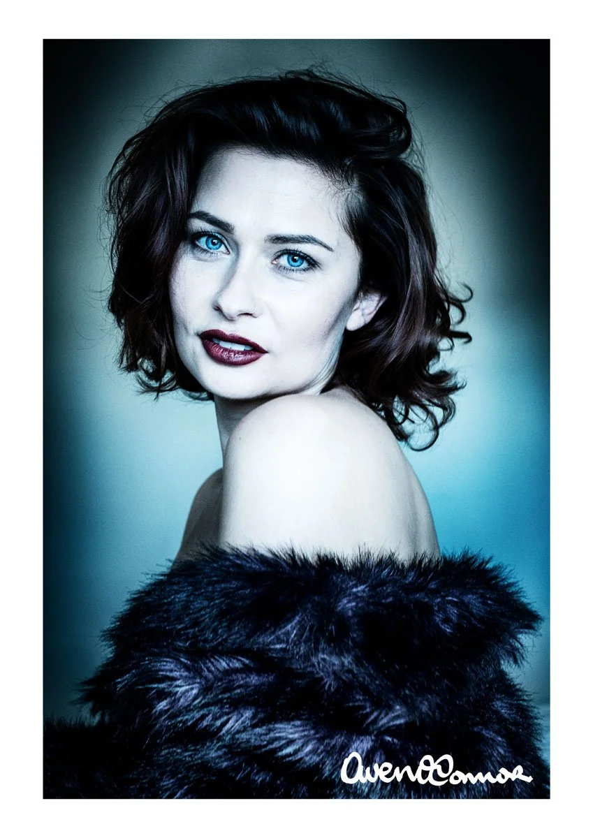 A woman with short, wavy dark hair and bright blue eyes, wearing dark lipstick and a black fur coat, standing against a gradient background.