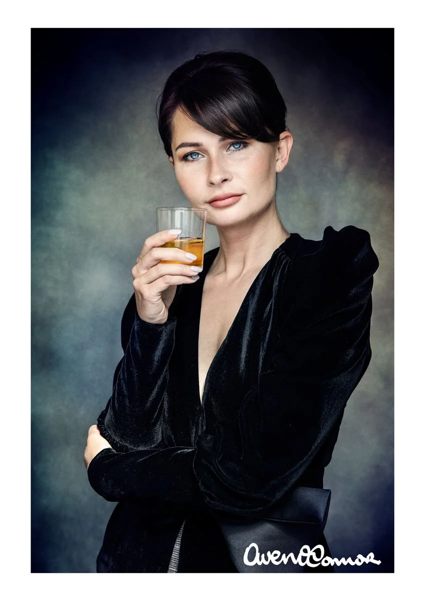 A woman with short dark hair and blue eyes holding a glass of whiskey, wearing a black velvet blazer, posing with a neutral background.