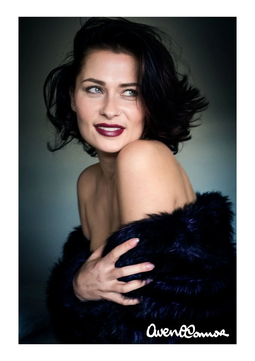A woman with short, dark, wavy hair and fair skin, wearing dark lipstick, looking to her left, wrapped in a black fur stole, against a dark background.