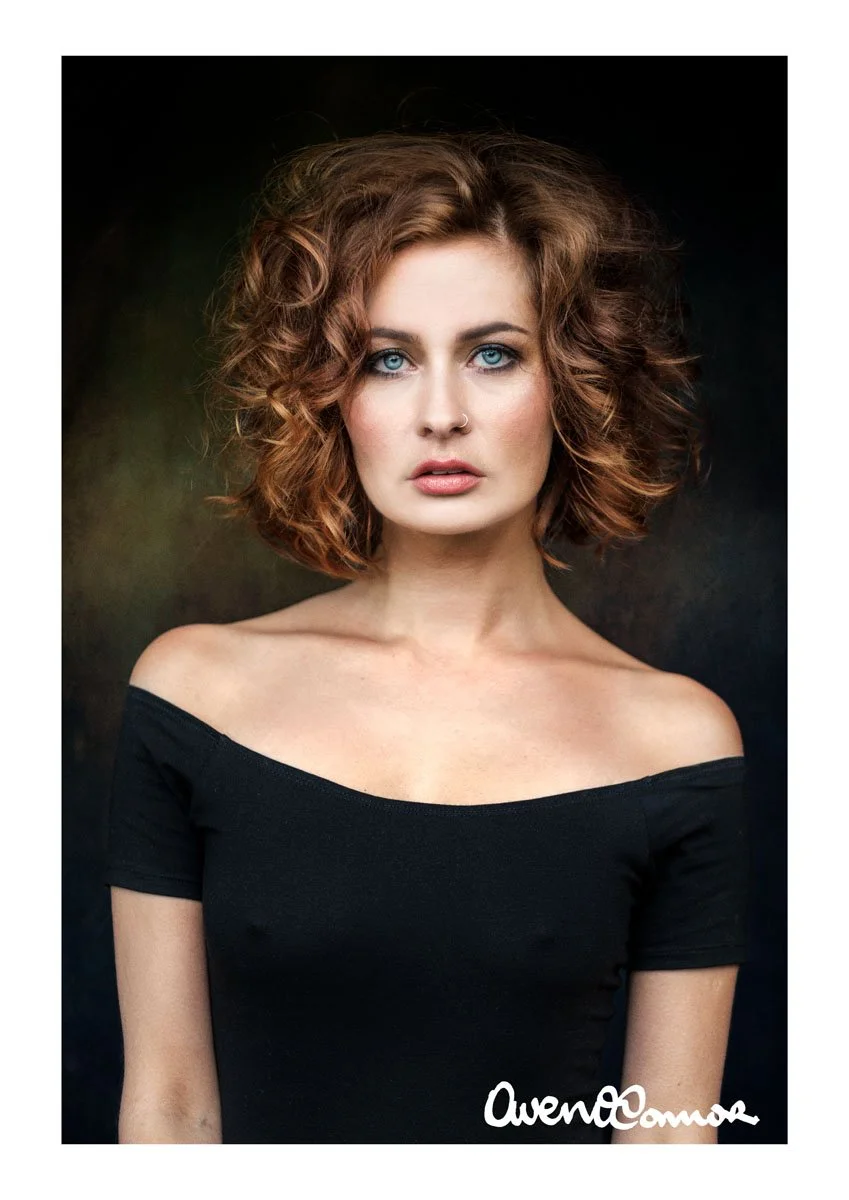 Portrait of a woman with curly auburn hair, blue eyes, and light skin, wearing a black off-the-shoulder top against a dark background.
