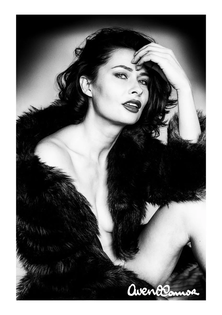 Black and white portrait of a woman with dark hair wearing a fur coat, with one hand on her forehead and gazing at the camera, signed 'Owen Cannon'.