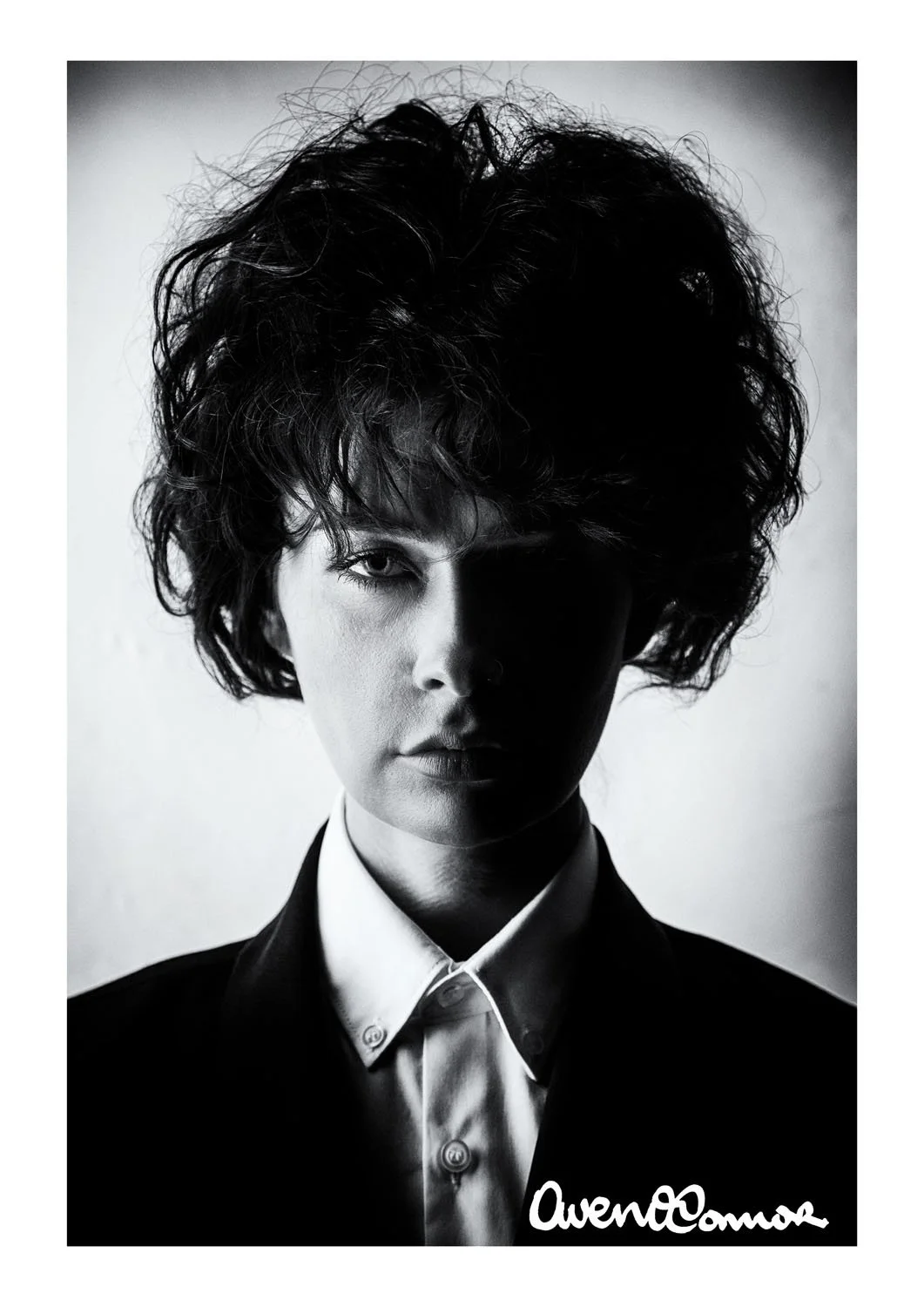Black and white portrait of a woman with curly hair, wearing a shirt and blazer, with one eye partially covered in shadow, and a signature reading 'Owen Cannon' at the bottom right.