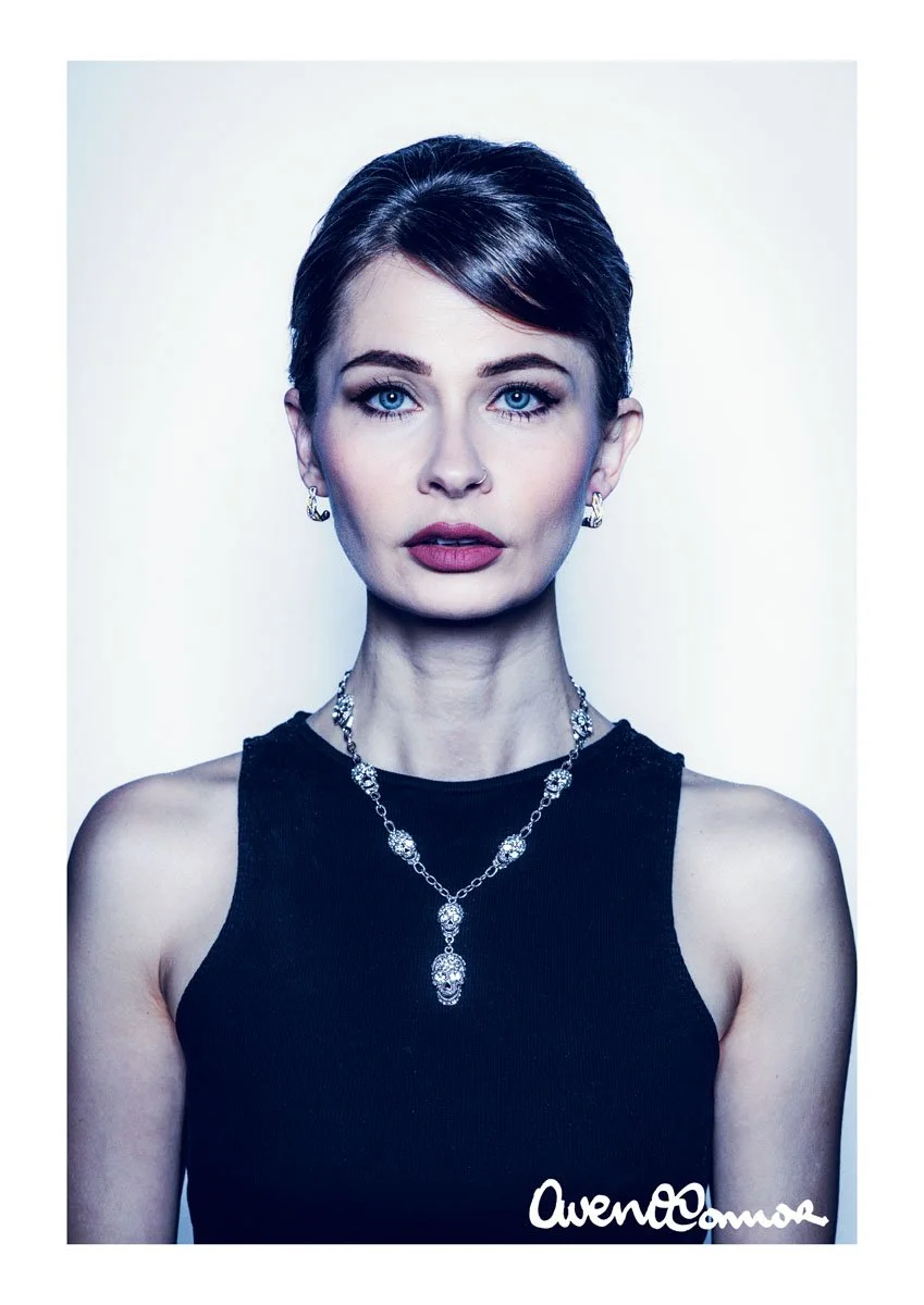 Portrait of a woman with short dark hair and blue eyes, wearing a black sleeveless top and silver jewelry, against a plain background.