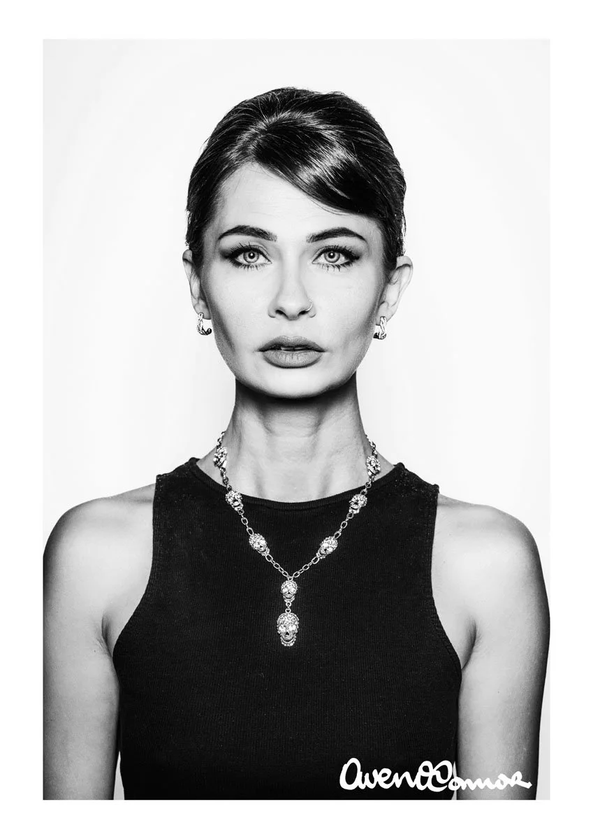 Black and white portrait of a woman with short dark hair, wearing a sleeveless top, earrings, and a necklace with ornate jewelry, in front of a plain background.