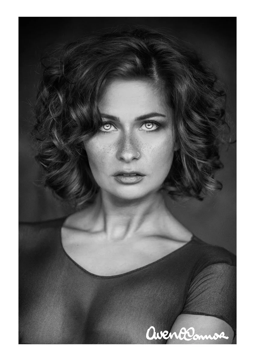 Black and white portrait of a woman with curly hair, intense eyes, a nose ring, wearing a sheer top, signature at the bottom right corner.