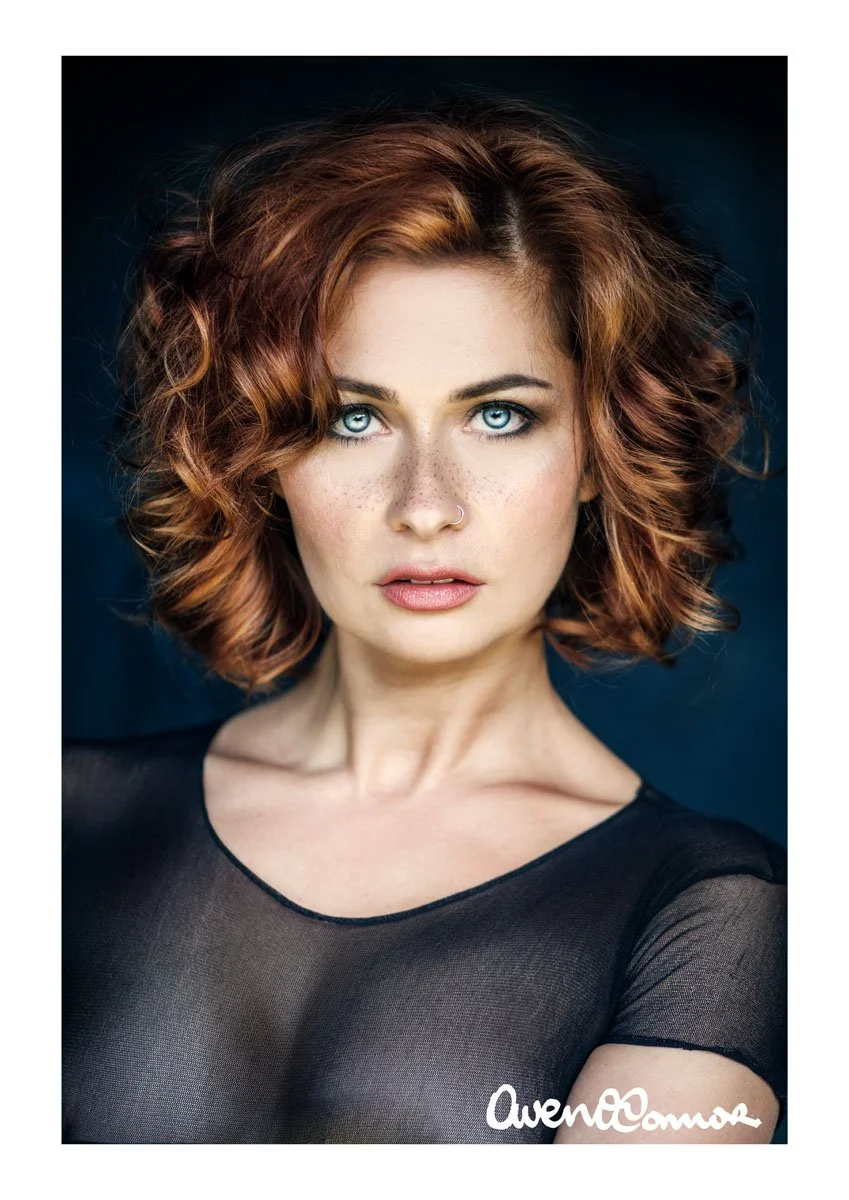 A woman with short, curly red hair and blue eyes, wearing a black, sheer top, looking directly at the camera with a confident expression.