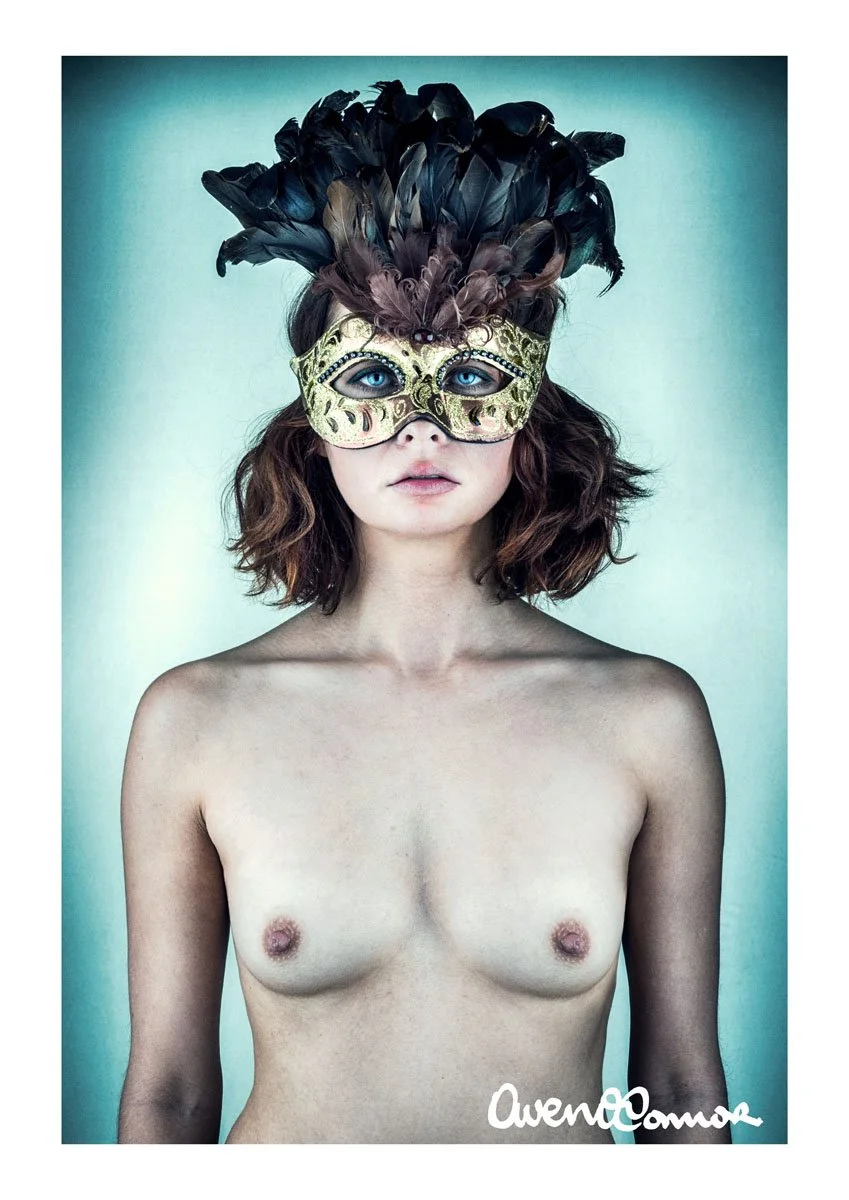 A topless woman with shoulder-length wavy brown hair wearing an ornate gold and black feathered mask with blue eyes visible through the mask. The background is plain and pale. The image appears artistic and stylized.