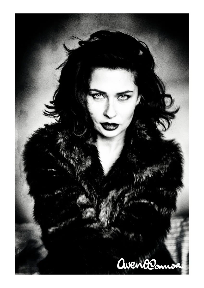 Black and white portrait of a woman with dark, wavy hair, piercing eyes, and bold lipstick, wearing a fur coat, with a signature at the bottom right corner.