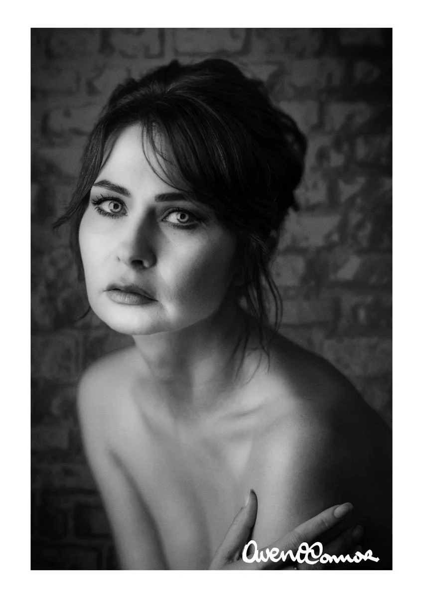 Black and white portrait of a woman with short dark hair, looking directly at the camera with light-colored eyes, set against a textured brick wall background.