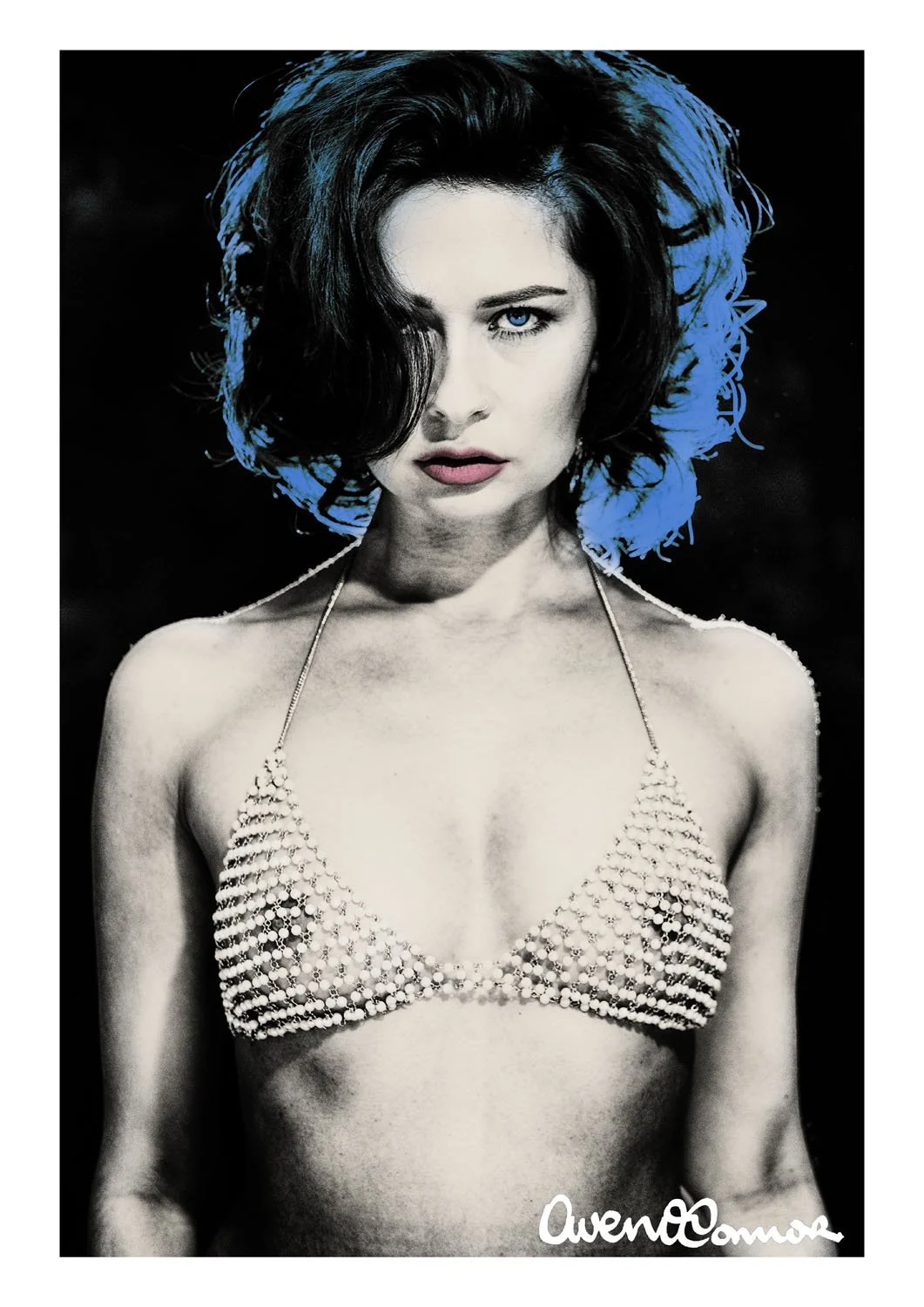 A black-and-white photograph of a woman with dark hair and blue eyes, wearing a pearl-studded bikini top, with a blue background and signature at the bottom right.