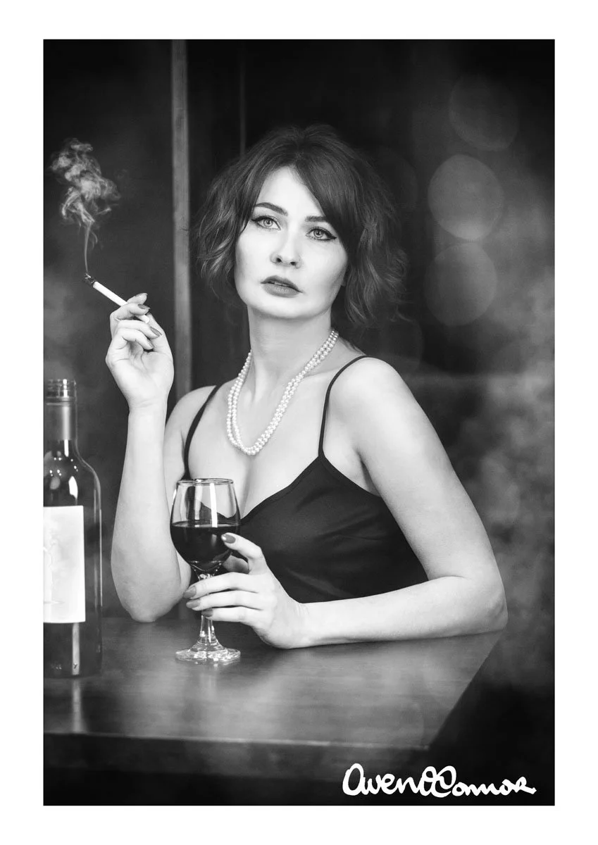 Black and white photo of a woman with short hair, wearing a black dress and pearl necklace, holding a glass of red wine and smoking a cigarette, with a bottle of wine on the table.