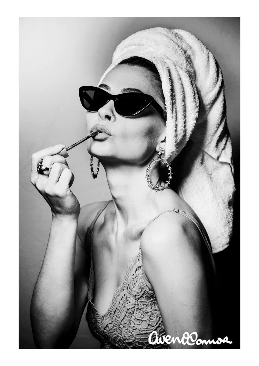 A black-and-white portrait of a woman wearing large sunglasses, earrings, and a lace top with a towel wrapped around her head. She is holding a lipstick applicator near her lips.