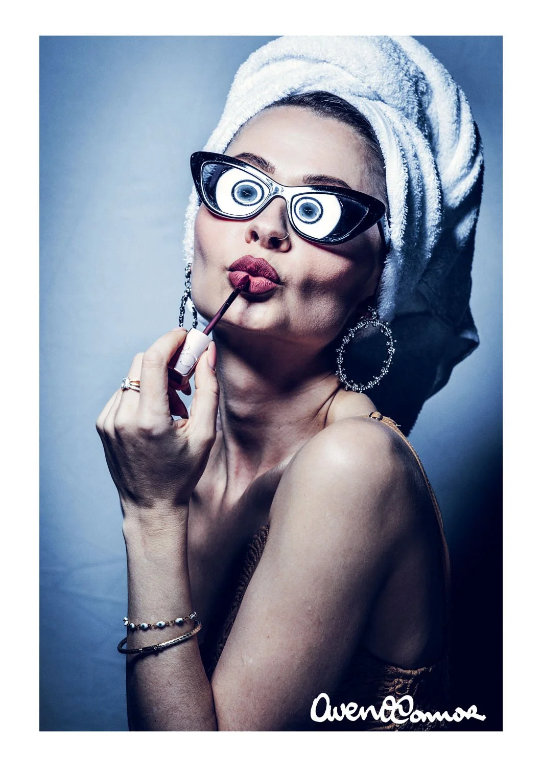 A woman is applying lipstick while wearing large sunglasses, a towel wrapped around her head, and jewelry, with a plain background.