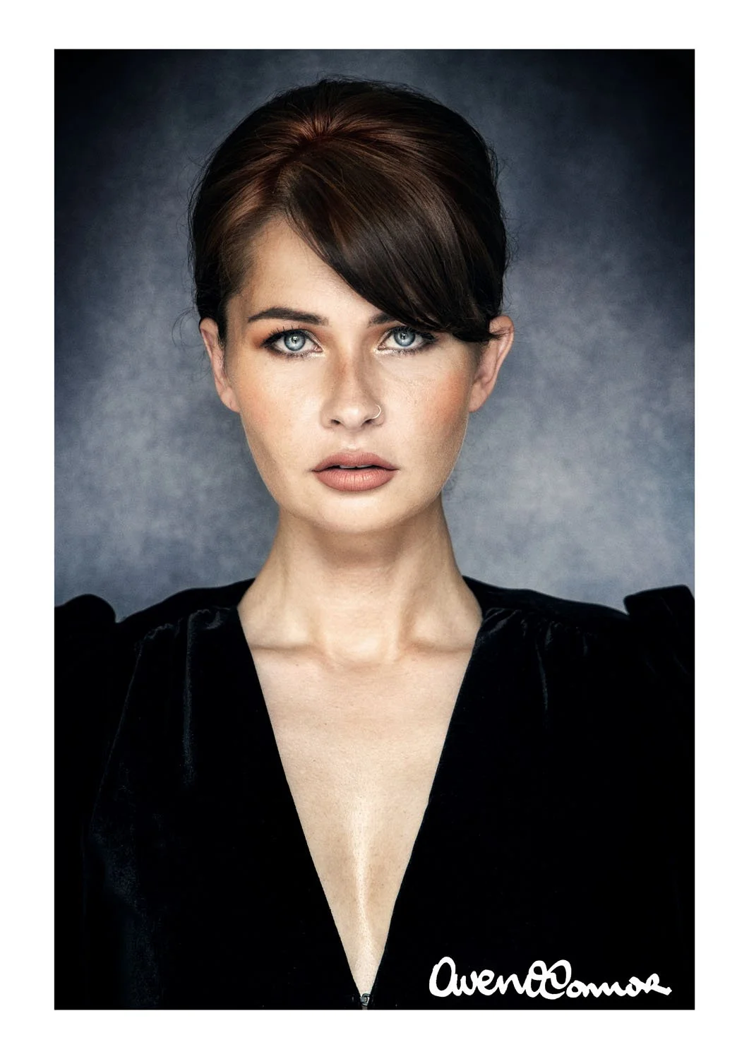 Portrait of a woman with dark brown hair, blue eyes, and light skin, wearing a black top, standing against a gray background.