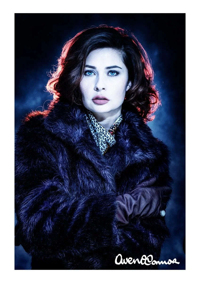 A woman with wavy brown hair and blue eyes wearing a black fur coat, a leopard print shirt, and brown gloves, posing against a dark background with dramatic lighting.