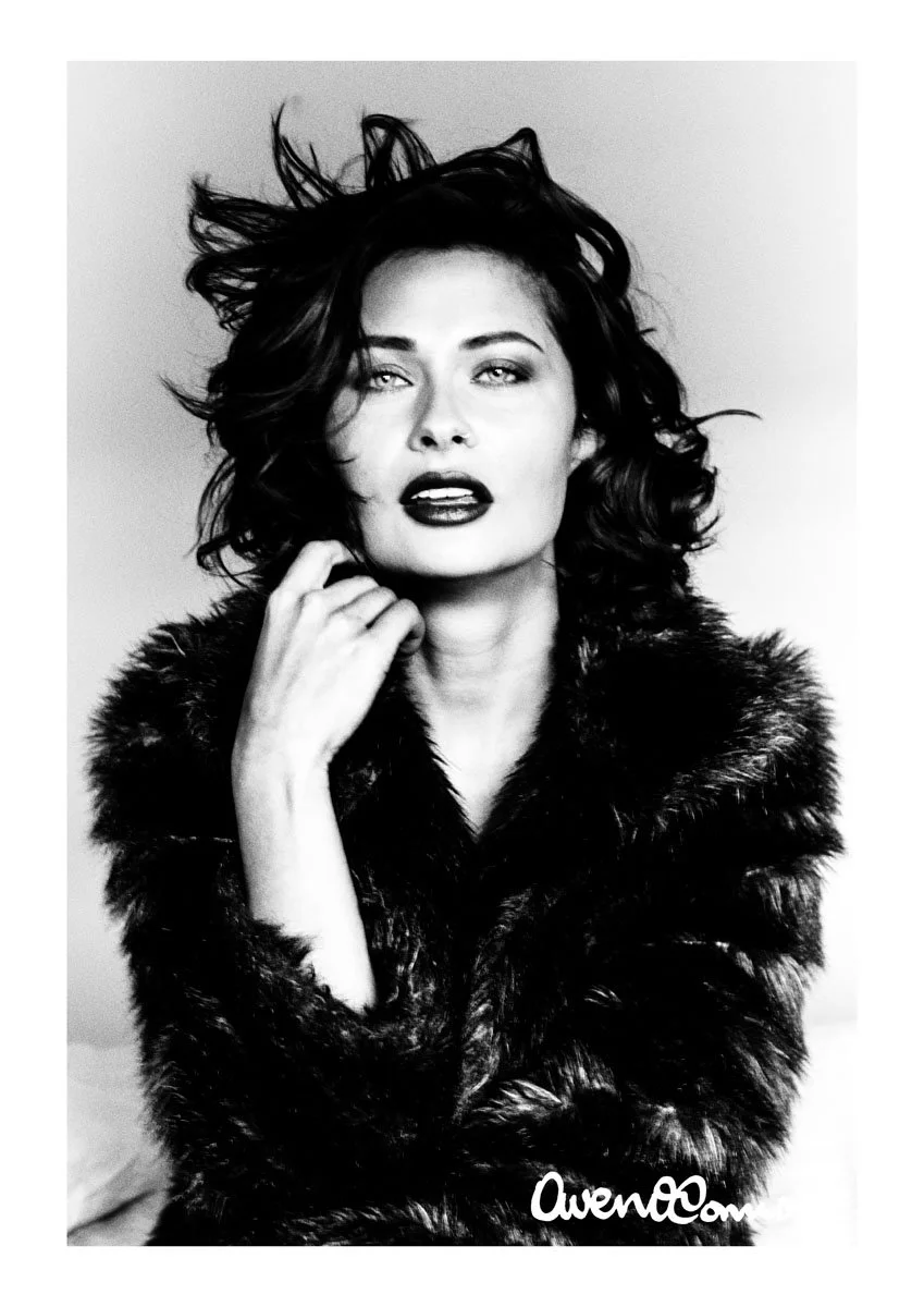 Black and white photo of a woman with curly hair, wearing a fur coat, looking at the camera with slightly parted lips, one hand near her face, in a studio setting.