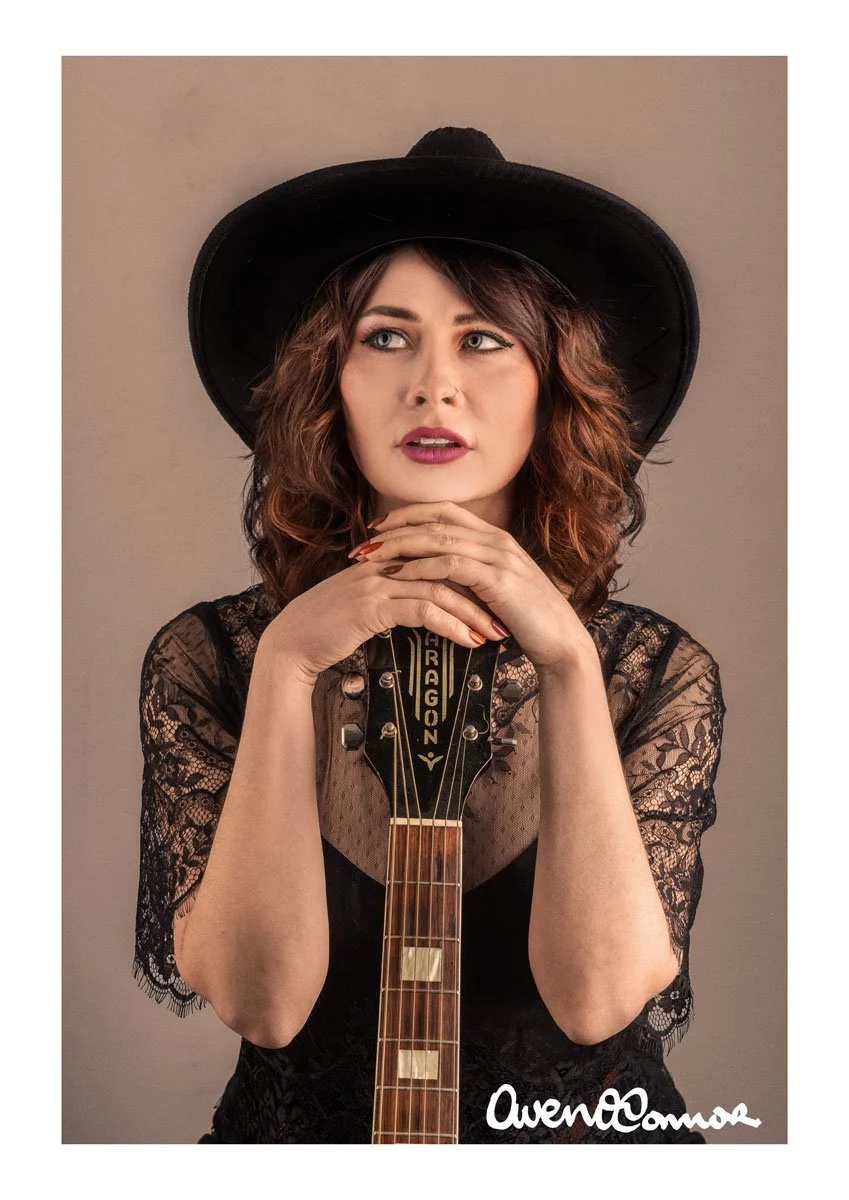 A woman with curly brown hair wearing a black wide-brimmed hat and black lace top, holding a guitar with the headstock showing. She has her hands clasped under her chin and looks to the side. The background is plain, and there is a signature at the b