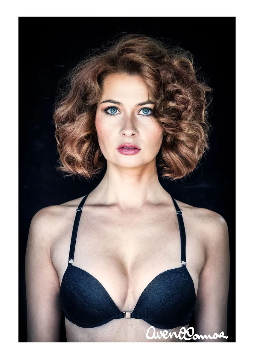 A woman with curly, shoulder-length red hair, blue eyes, and fair skin, wearing a black bra with thin straps against a dark background.