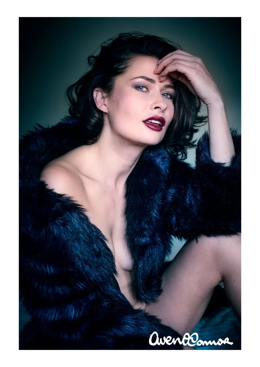 A woman with dark hair, light makeup, and deep red lipstick, wearing a dark fur coat, looking at the camera with her hand touching her forehead.