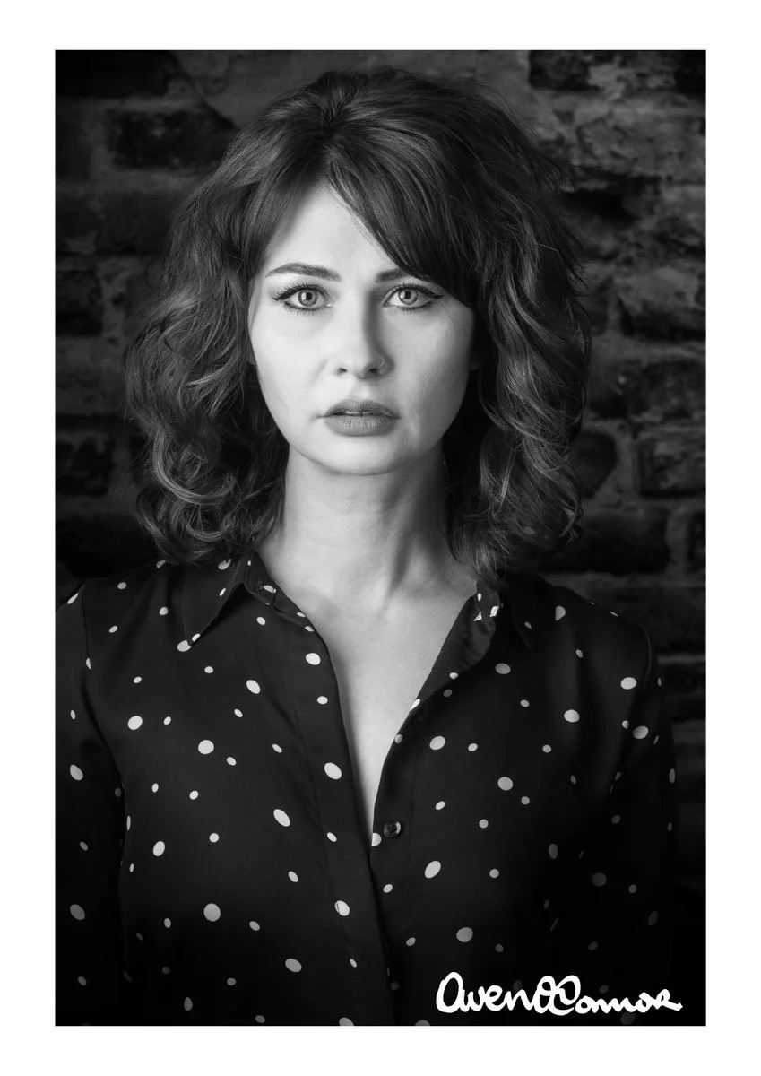 Black and white portrait of a woman with wavy hair, wearing a polka dot shirt, standing against a brick wall background.