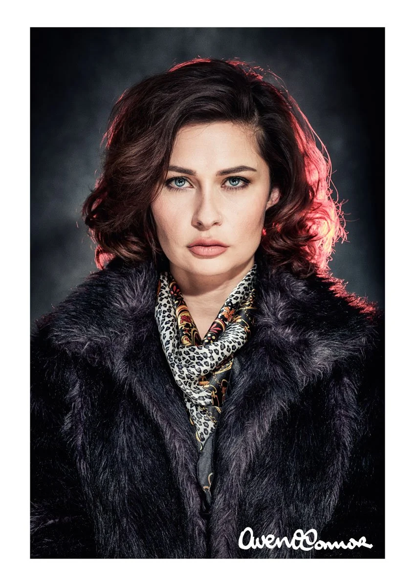 Portrait of a woman with wavy dark red hair, wearing a black fur coat and a leopard print scarf with gold accents, against a dark background with subtle lighting.