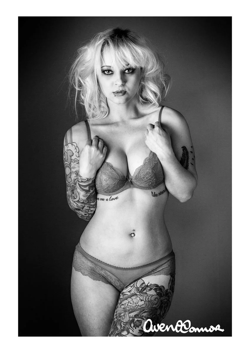 Black and white photo of a woman with tattoos in underwear, holding the straps of her bra, with messy blonde hair, and intense eye makeup. The photo is signed with the name 'AvenOcom' at the bottom right corner.