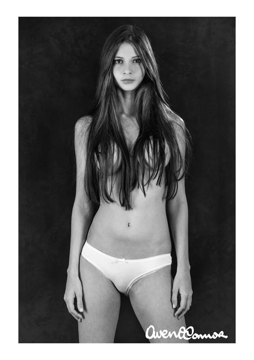 Black and white photo of a woman with long hair, wearing only underwear, standing against a dark background. Signed by Owen Moore.