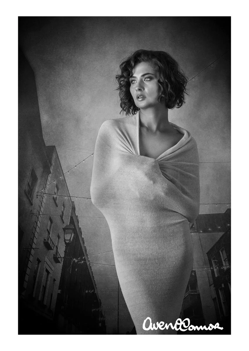 Black and white portrait of a woman with curly hair, wearing an oversized sweater, standing outside in an urban area with buildings and streetlights in the background.