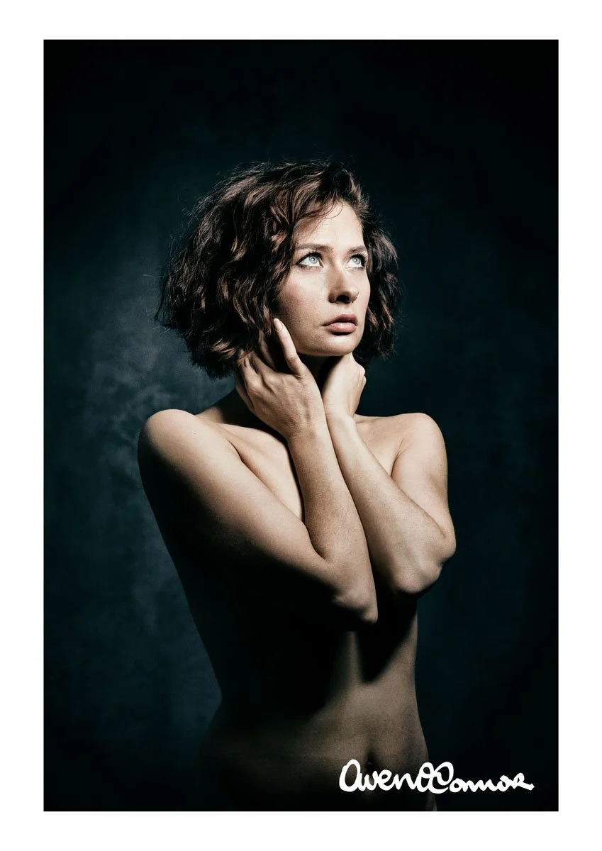 A young woman with curly brown hair, looking upward with a thoughtful expression, holding her neck with one hand and crossing her arms over her chest, against a dark background.
