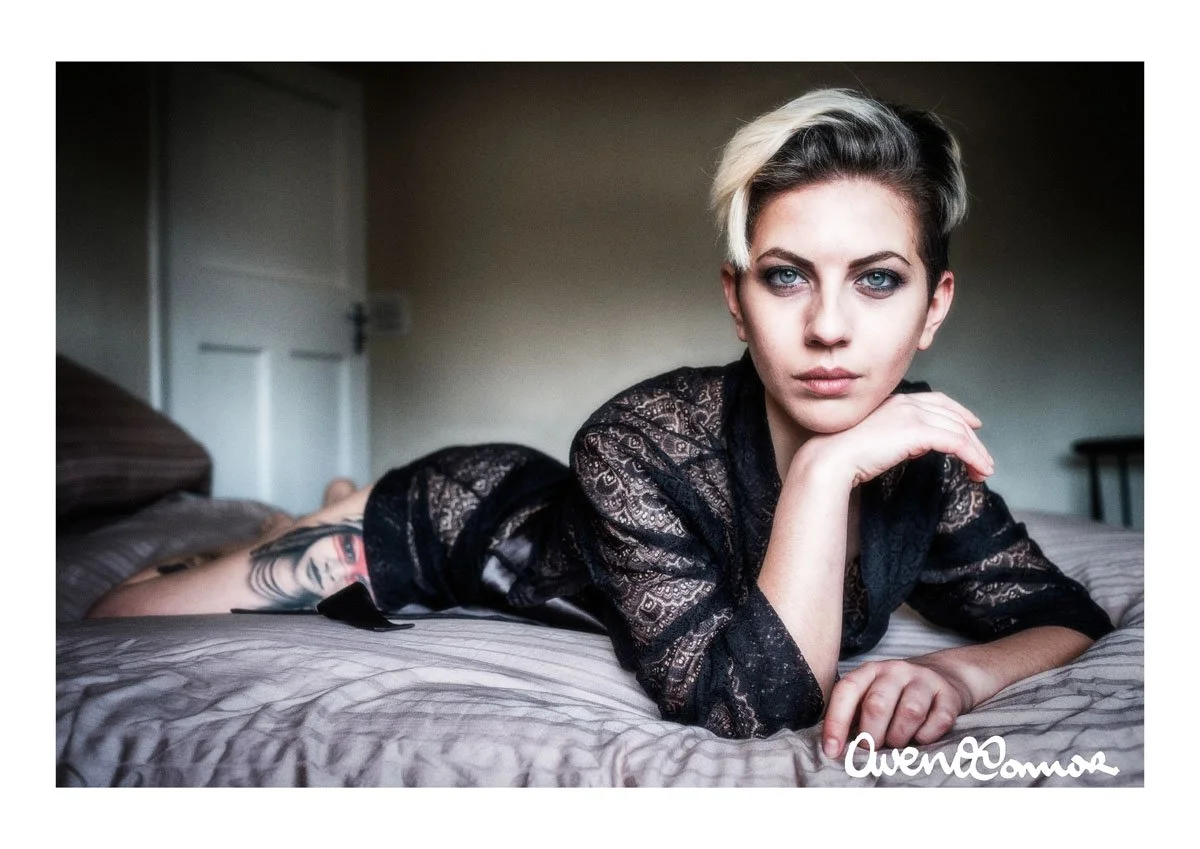 A woman with short blonde and dark hair lying on her stomach on a bed, wearing a black lace top, with a tattoo visible on her left arm. She has her chin resting on her left hand and is looking at the camera.