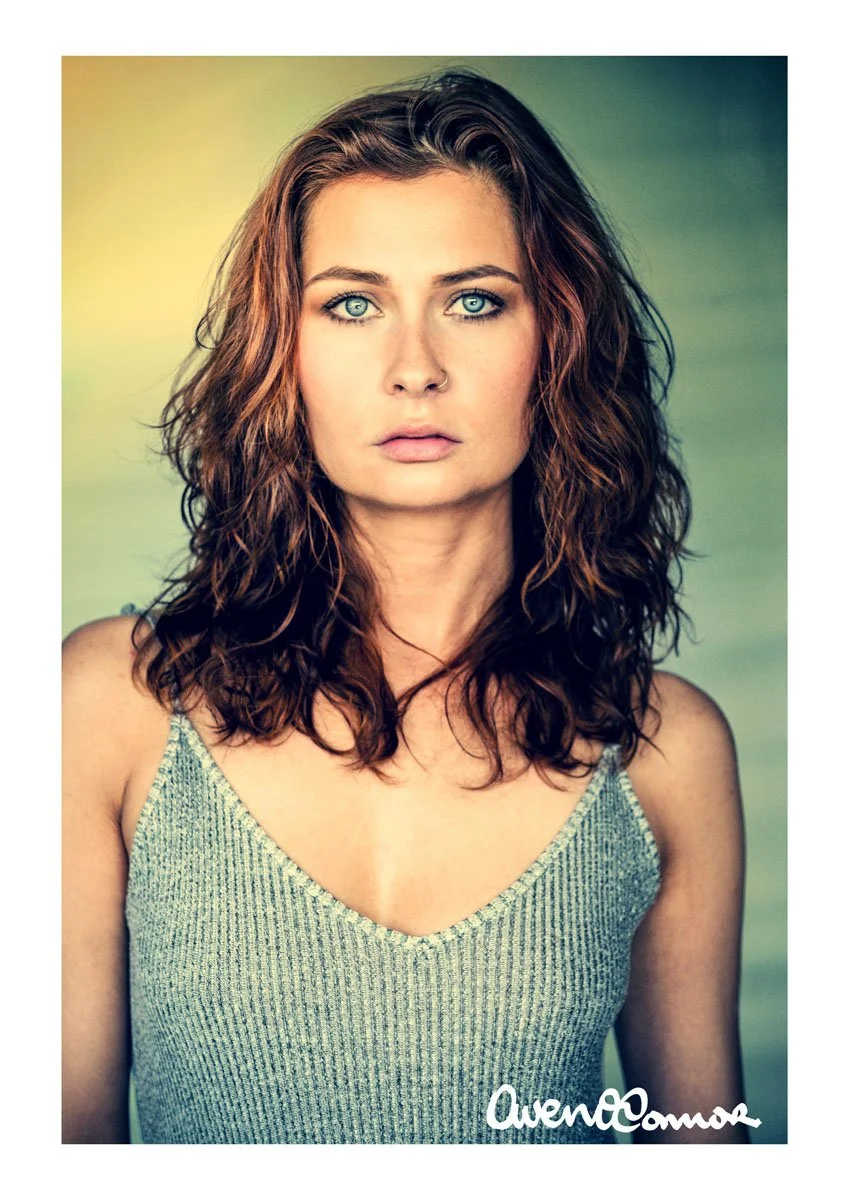 A portrait of a woman with wavy, shoulder-length brown hair, blue eyes, fair skin, wearing a sleeveless, V-neck, gray and white striped top. She has a neutral expression and is looking directly at the camera, with a blurred greenish background.