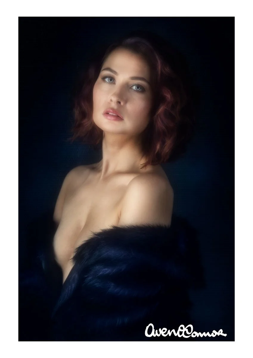 Portrait of a woman with wavy auburn hair, blue eyes, and fair skin, wearing a black fur coat off her shoulders against a dark background.